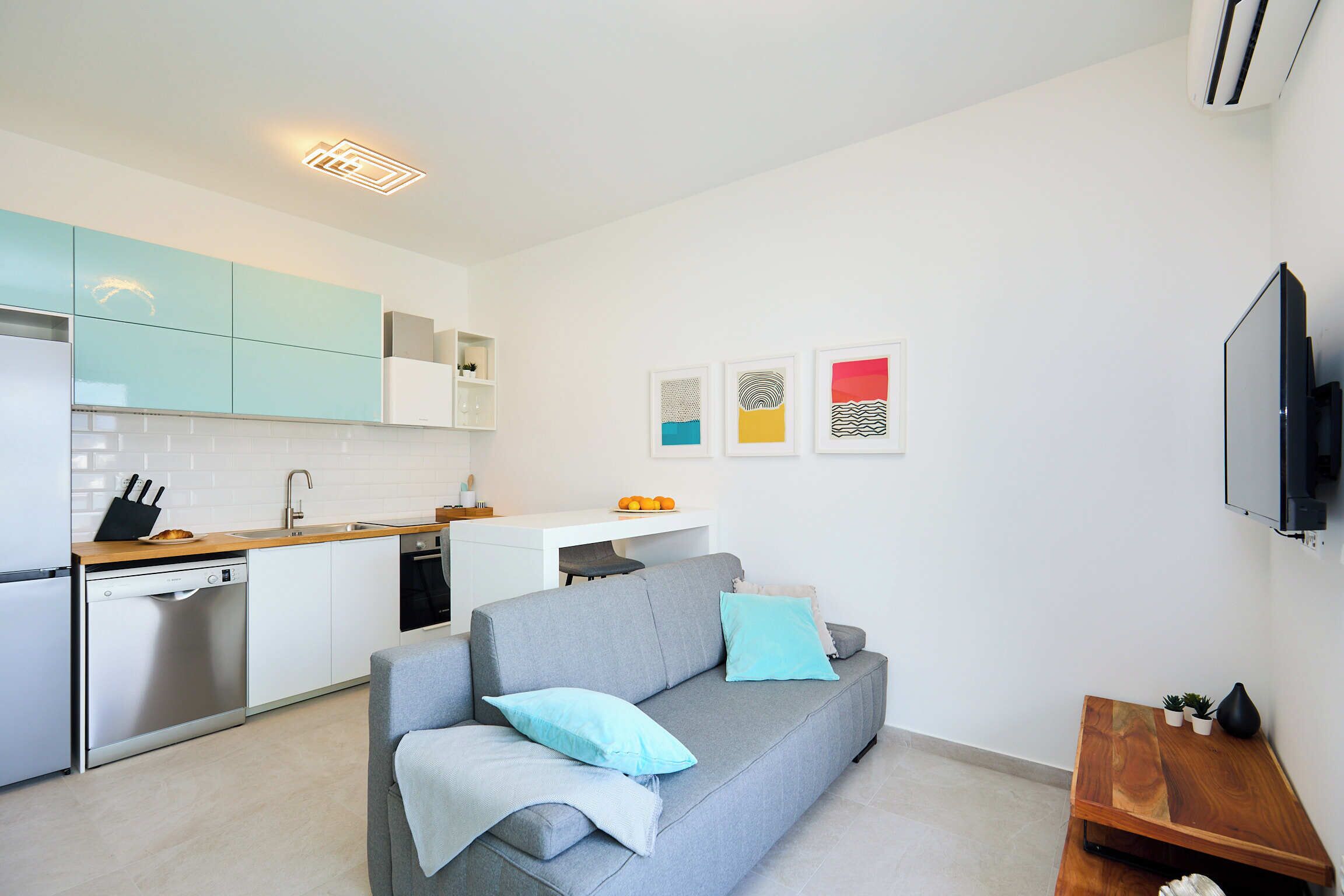 A modern apartment with a grey sofa, kitchen area, and a flat-screen TV.