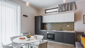 A modern kitchen with white cabinets, a black refrigerator, and a patterned backsplash. There is a dining table with chairs and a small kitchen island with a bowl of fruit and a book. - Venuto Home by Wonderful Italy - Deep Blue (Catania)