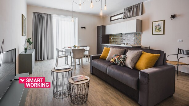 A modern living room with a sofa, coffee table, and TV. There is a kitchen area with cabinets and a window. - Venuto Home by Wonderful Italy - Deep Blue (Catania)