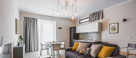 A modern living room with a sofa, coffee table, and TV. There is a kitchen area with cabinets and a window.