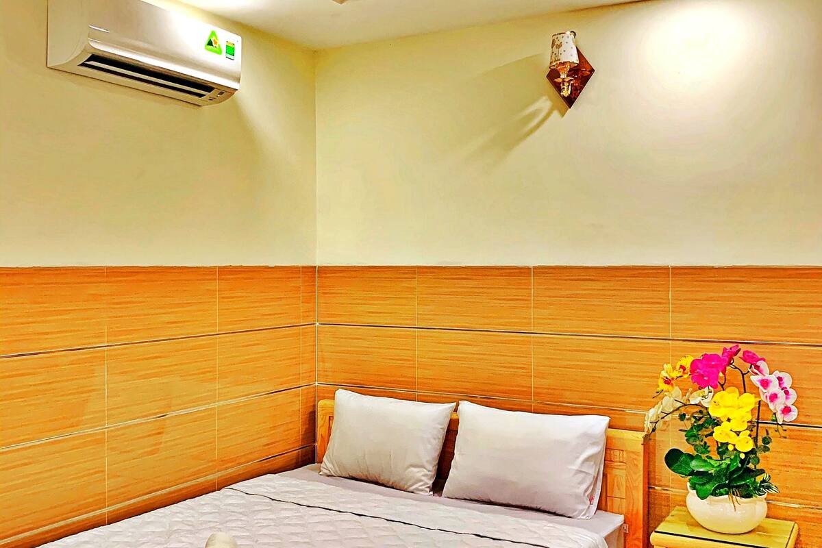 Een hotelkamer met een bed, een nachtkastje met een bloevas, een airconditioning en een wandlamp.