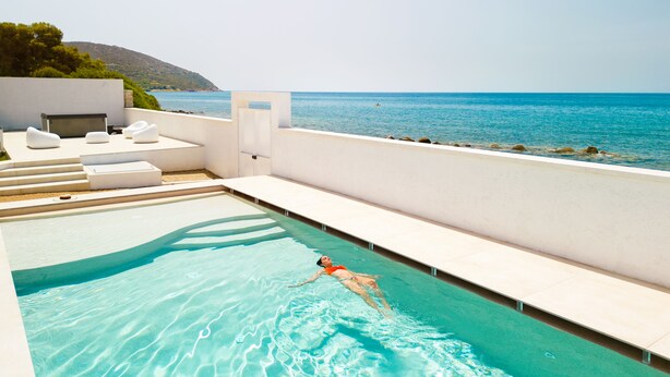Area piscina con una persona che nuota, solarium con divani bianchi e vista sul mare.