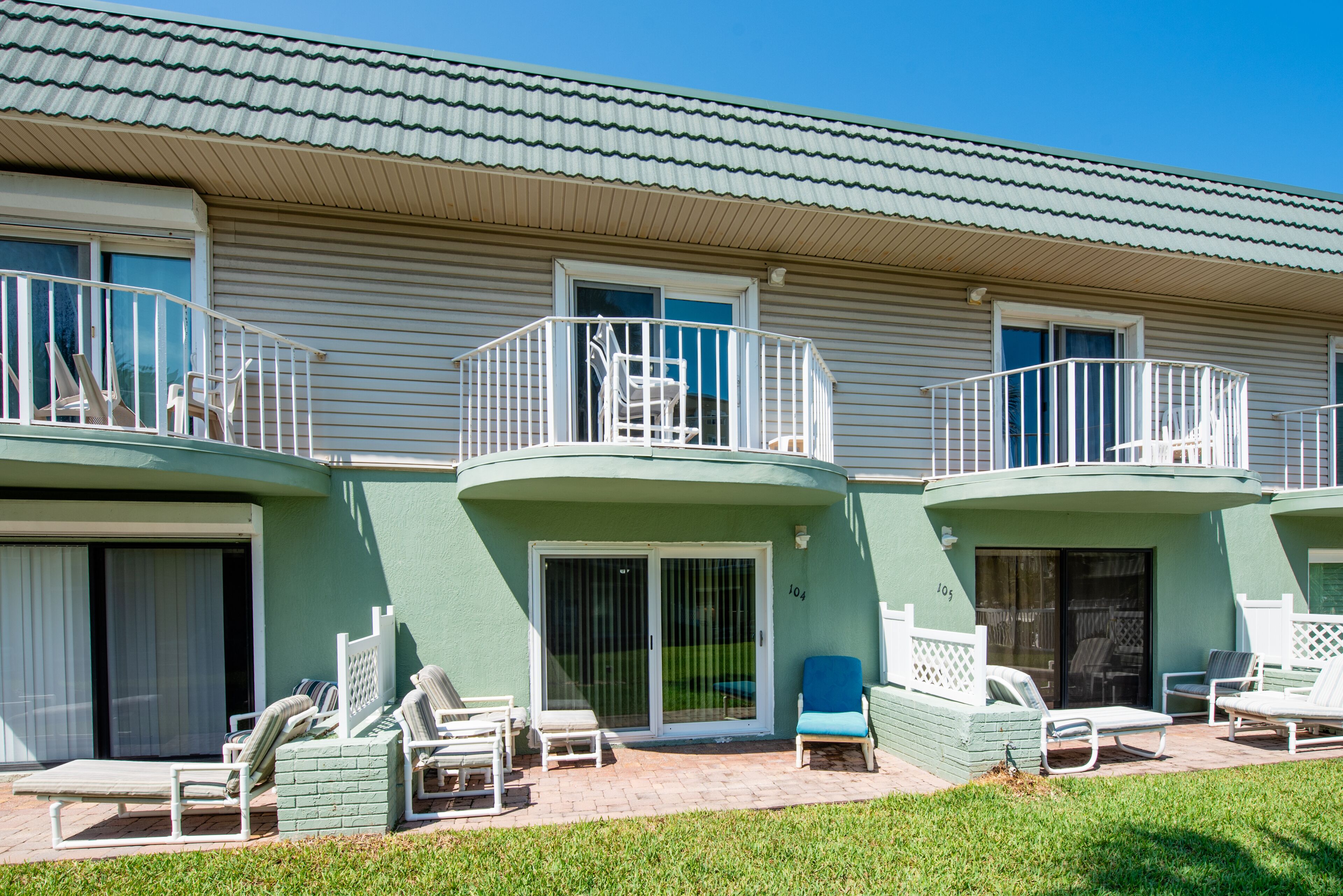 A two-story building with a green exterior, a balcony with white railings, and a patio area with lounge chairs and a small table.