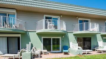 A two-story building with a green exterior, a balcony with white railings, and a patio area with lounge chairs and a small table.
