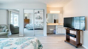 A bedroom with a bed, a chair, a TV stand, and a sliding door leading to a bathroom. - Balcony & A Patio, Private Beach Access, Heated Pool (Indialantic)