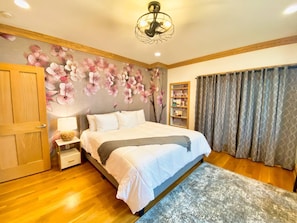 Un dormitorio con cama, suelo de madera, papel tapiz floral, una estantería y una ventana con cortinas.