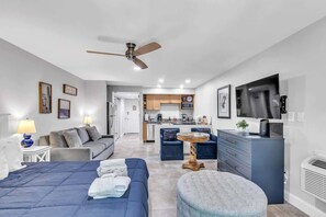 A modern hotel room with a bed, a sofa, a dining table, a TV, and a kitchenette.