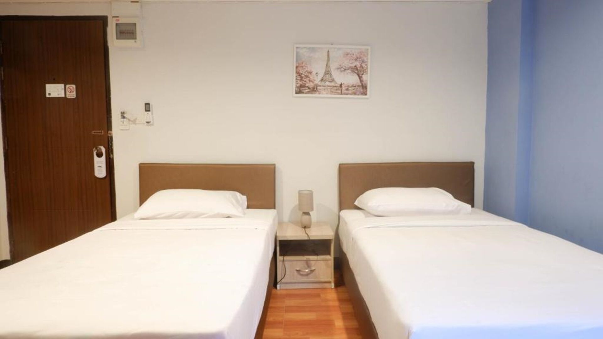 A hotel room with two single beds, a wooden door, a small bedside table with a lamp, and a framed picture of the Eiffel Tower on the wall.