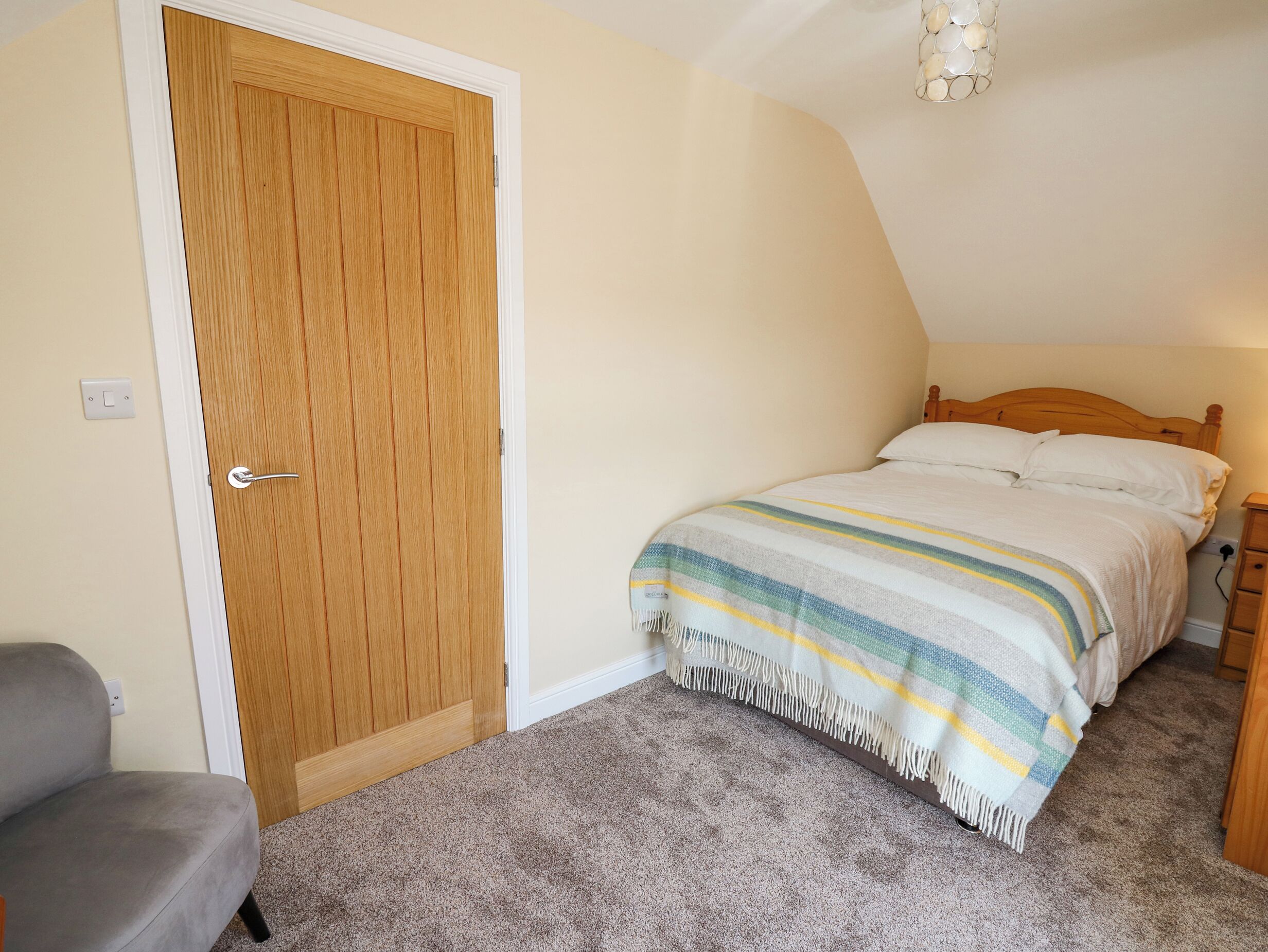 A bedroom with a wooden door, a bed with a striped blanket, a gray chair, and a carpeted floor.