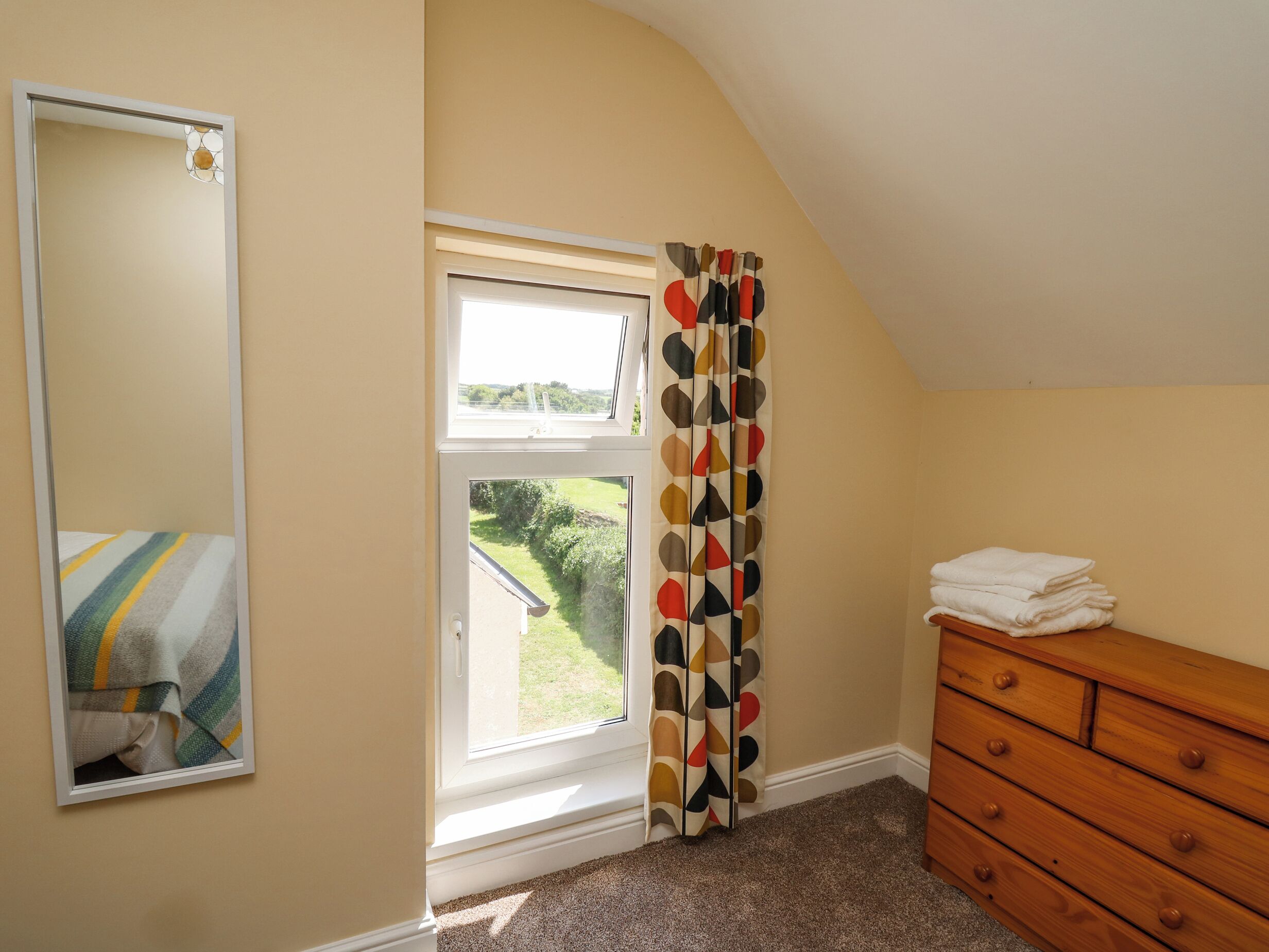 A bedroom with a large window, a mirror, a wooden dresser, and a colorful curtain.