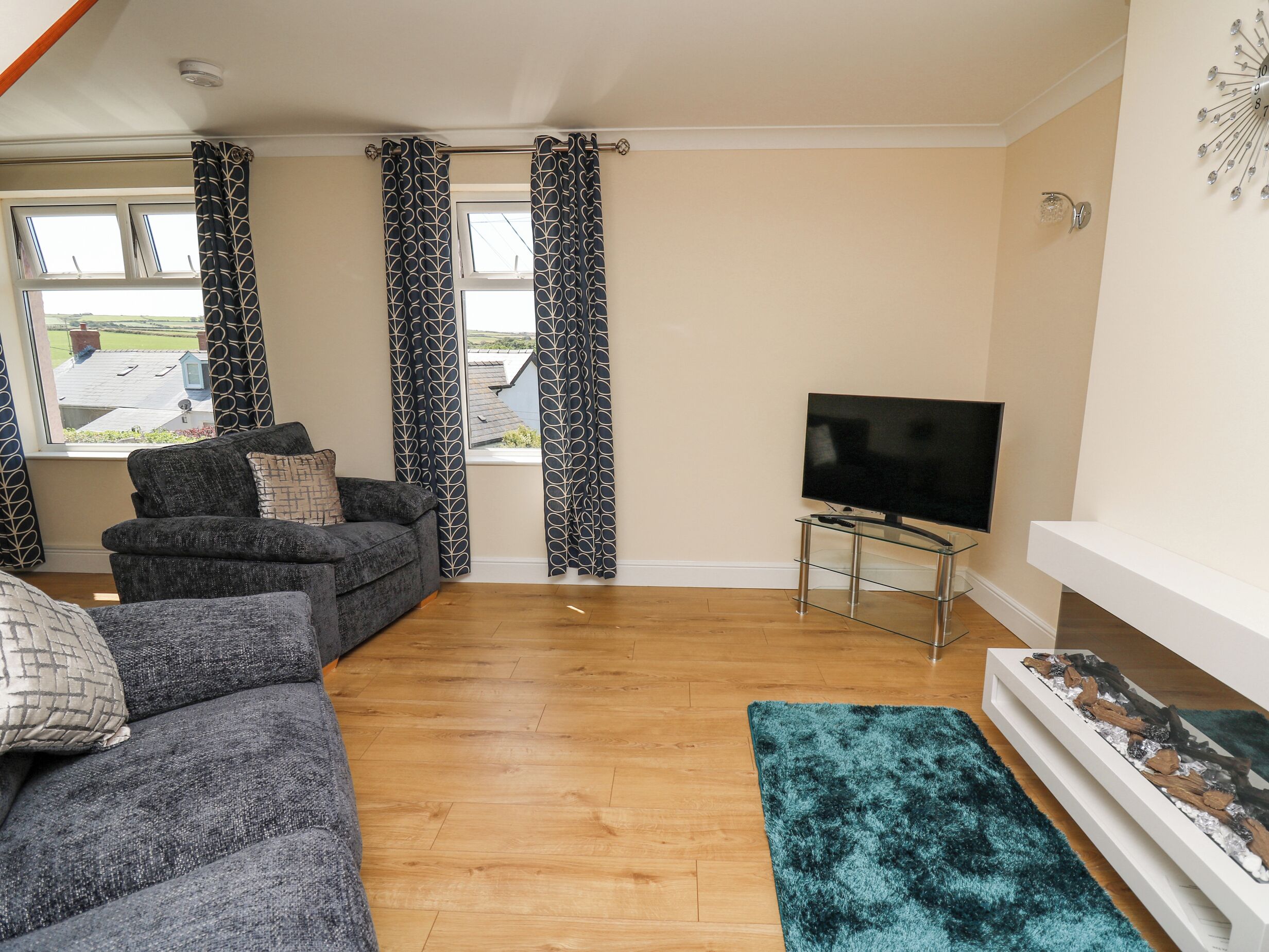A living room with a grey sofa, a flat-screen TV on a glass stand, a blue rug, and a fireplace with a decorative piece.