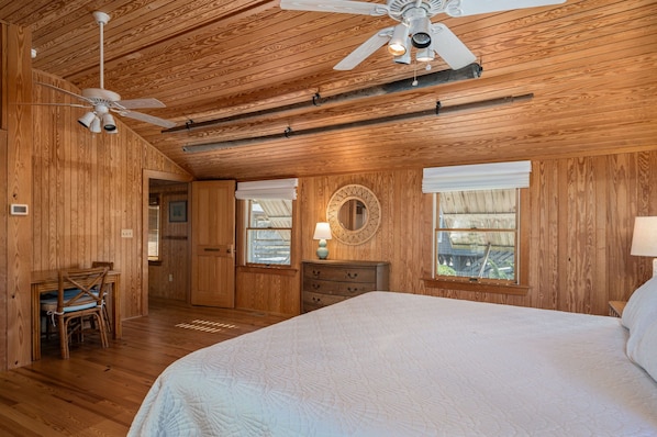 Un dormitorio con paneles de madera, una cama, un aparador, una silla, un ventilador de techo y una ventana con vistas.
