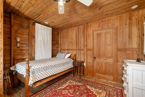 Une chambre à coucher aux murs lambrés, avec un lit, une table de chevet, une commode et un ventilateur de plafond.