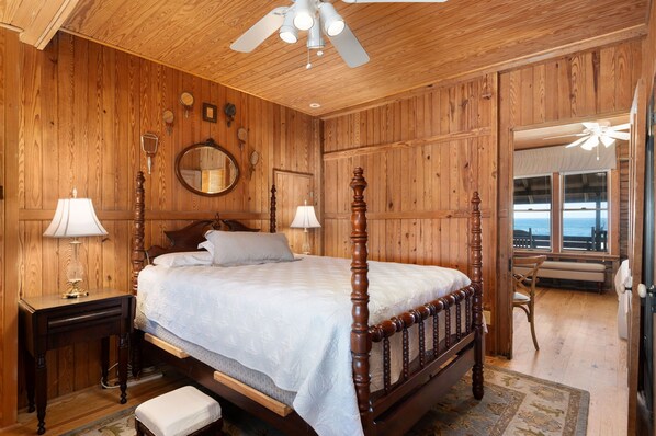 Une chambre à coucher aux murs lambrissés, dotée d’un lit à baldaquin, de deux lampes de chevet, d’un miroir et offrant une vue sur l’océan grâce à de grandes fenêtres.