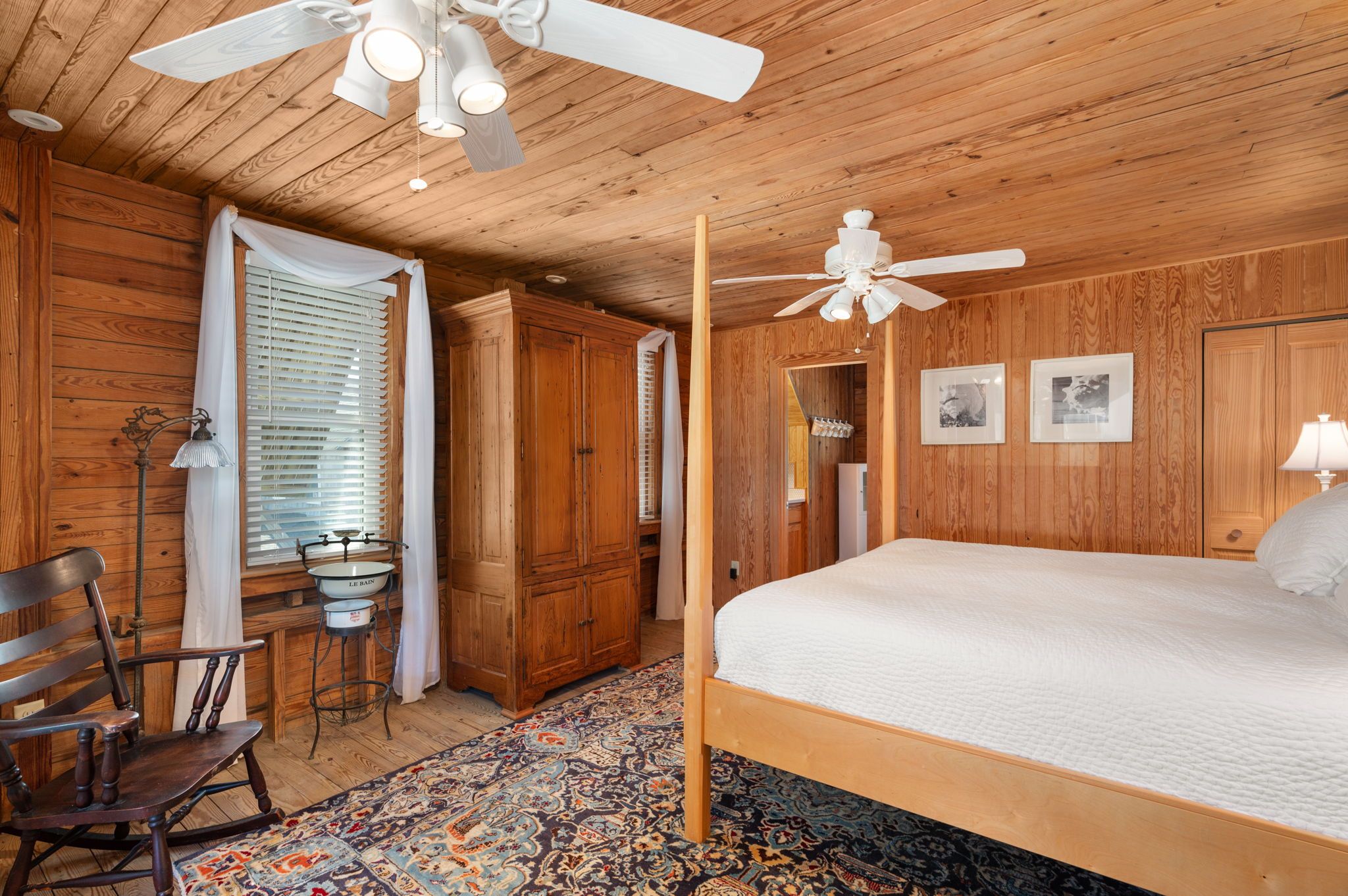 A wooden-paneled bedroom with a bed, a rocking chair, a ceiling fan, and a patterned rug.
