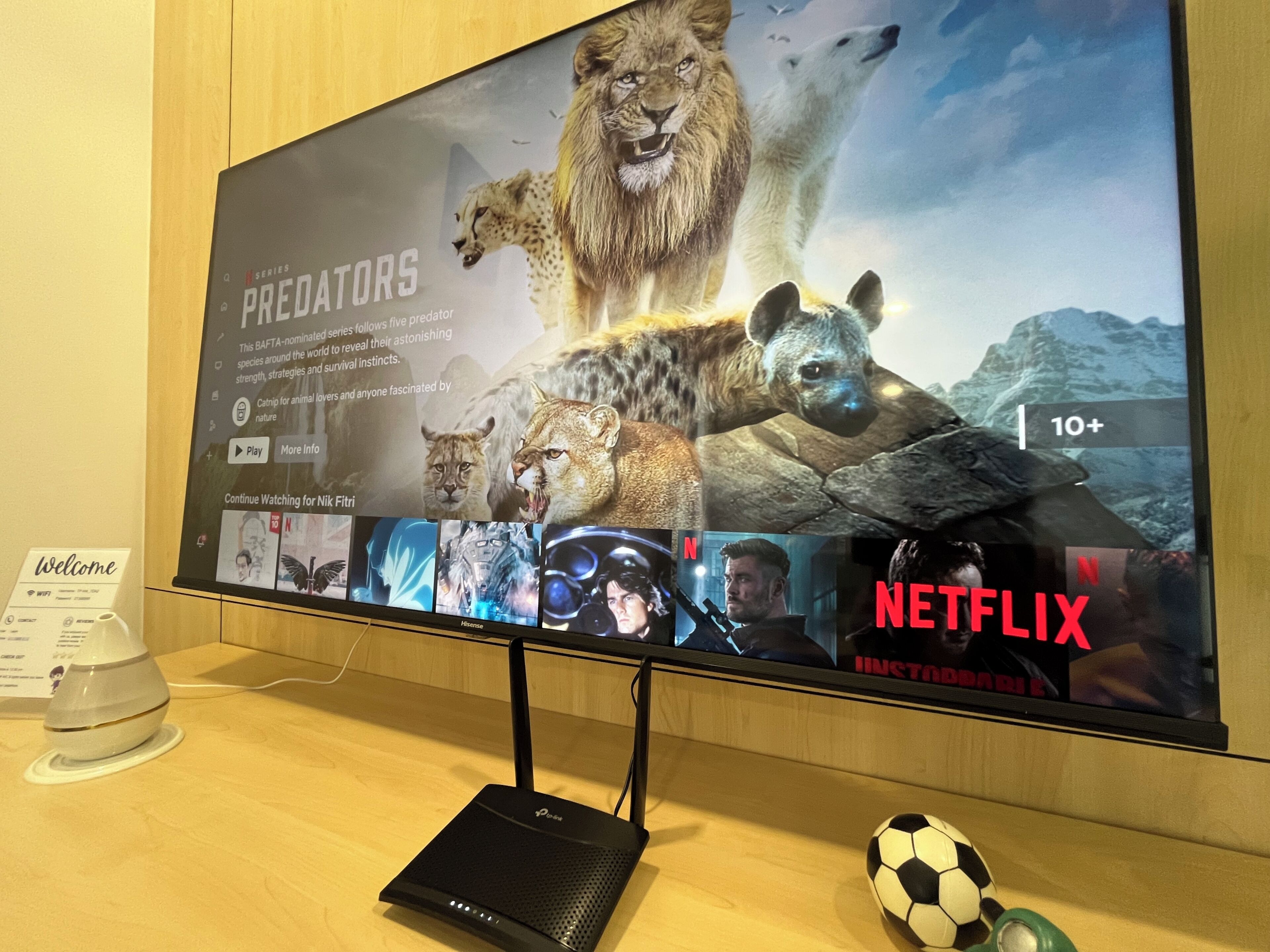 Un écran de télévision affiche une série de Netflix intitulée « Predators », avec une classification d’âge de « 10 ans et plus », un symbole Wi-Fi et un routeur portable noir posé sur une surface en bois.