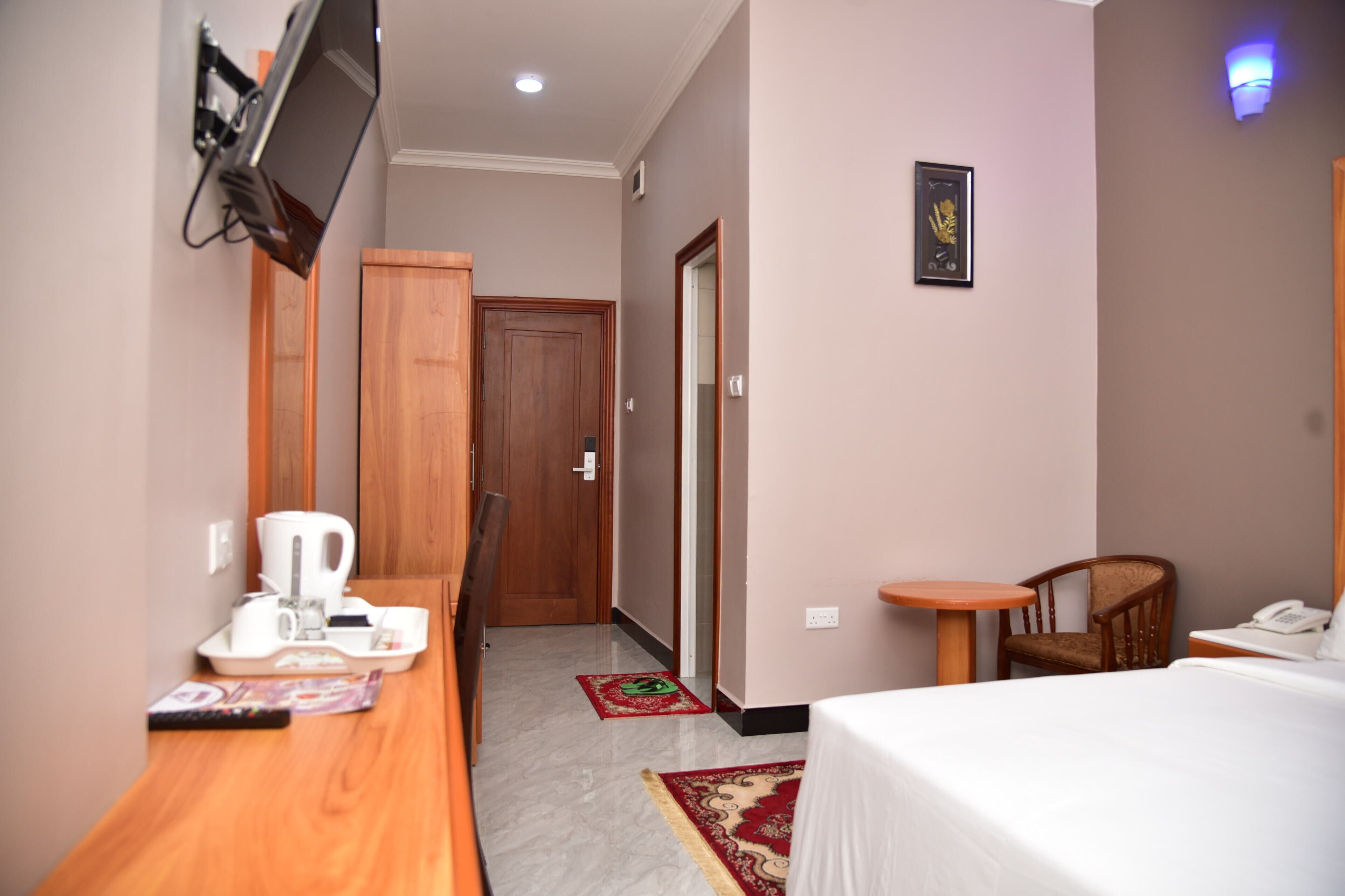 A hotel room with a bed, a desk with a coffee maker, a chair, a small table, a TV mounted on the wall, and a door leading to another room.
