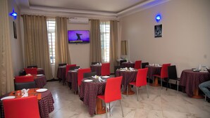Un comedor con varias mesas y sillas, un televisor montado en la pared y una ventana con cortinas.