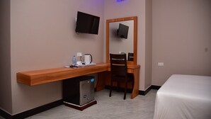 Habitación de hotel con escritorio de madera, una silla, una cama, un refrigerador pequeño, un televisor montado en la pared y un espejo sobre el escritorio.