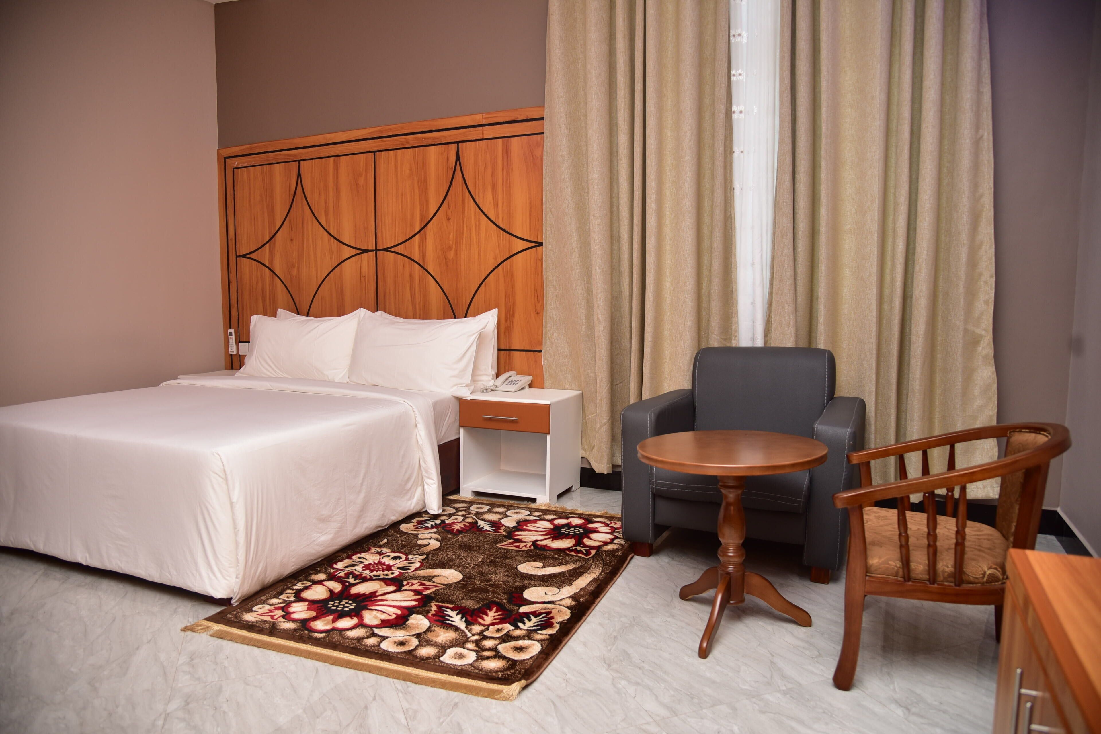 Una habitación de hotel con cama, una mesita, una silla, una mesita de noche y un cabecero de madera.