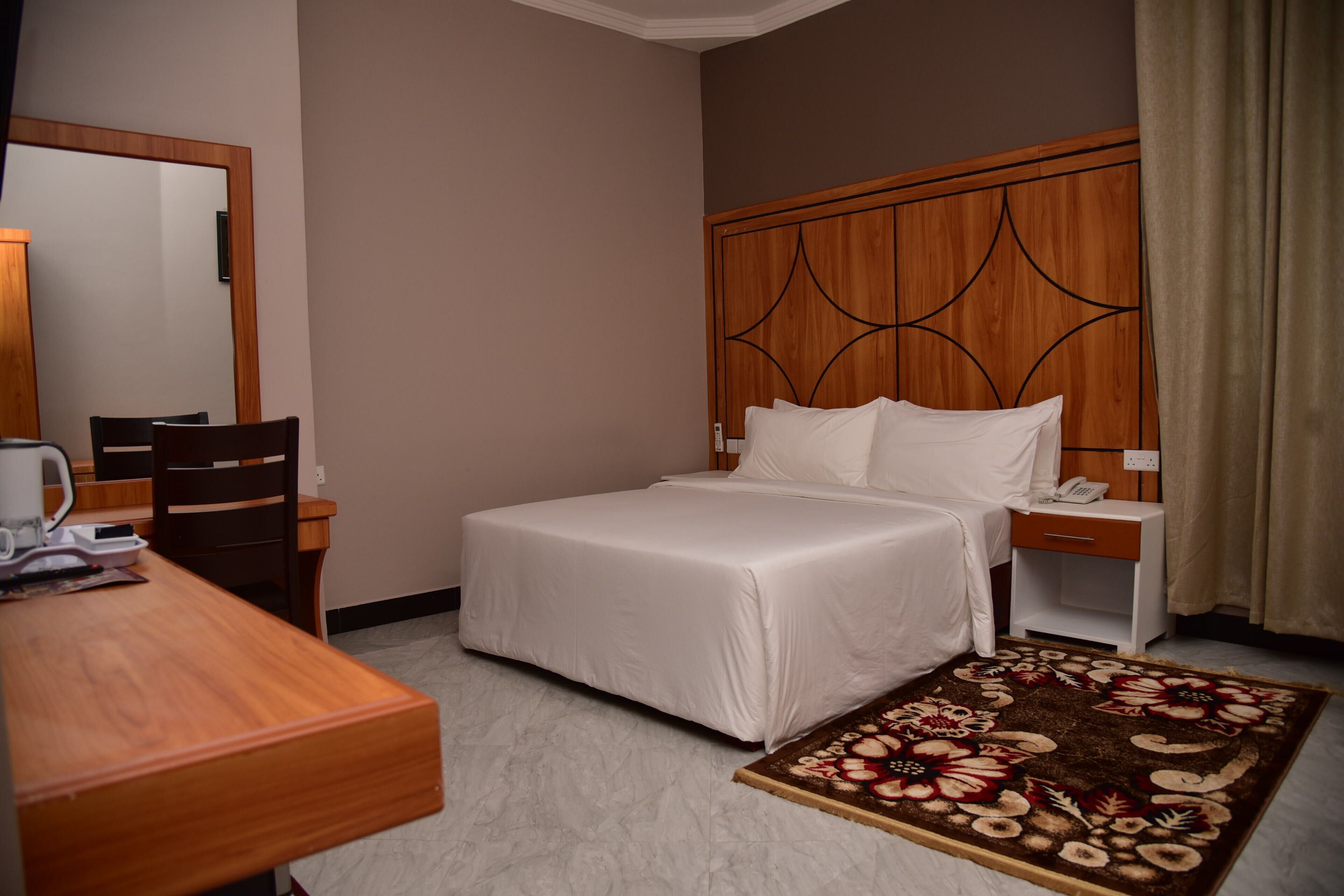 A hotel room with a bed, a desk, a chair, a mirror, and a patterned rug.
