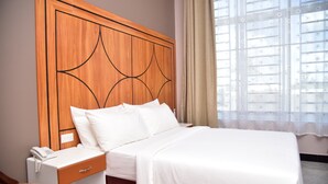 Habitación de hotel con cabecera de madera, una cama individual con ropa blanca, una mesita de noche con teléfono y una ventana con cortinas traslúcidas.