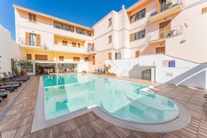 A pool area with lounge chairs, a covered seating area, and a building with multiple balconies.