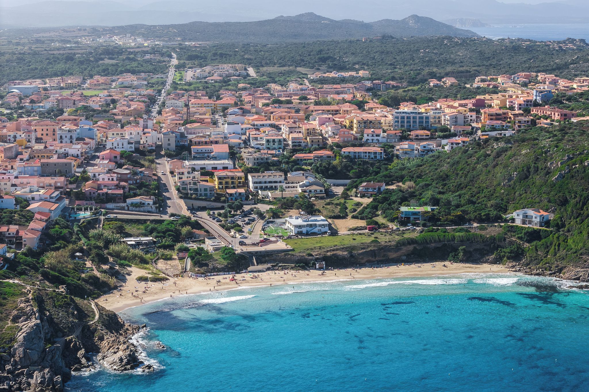 An aerial view of a coastal town with a beach, buildings, and a mountainous backdrop.