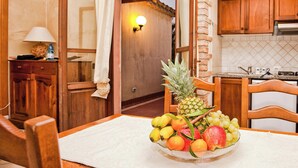 A room with a dining table set with fruit, wooden cabinets, and a kitchen area with a stove. - Residence Baia Delle Palme (Pula)