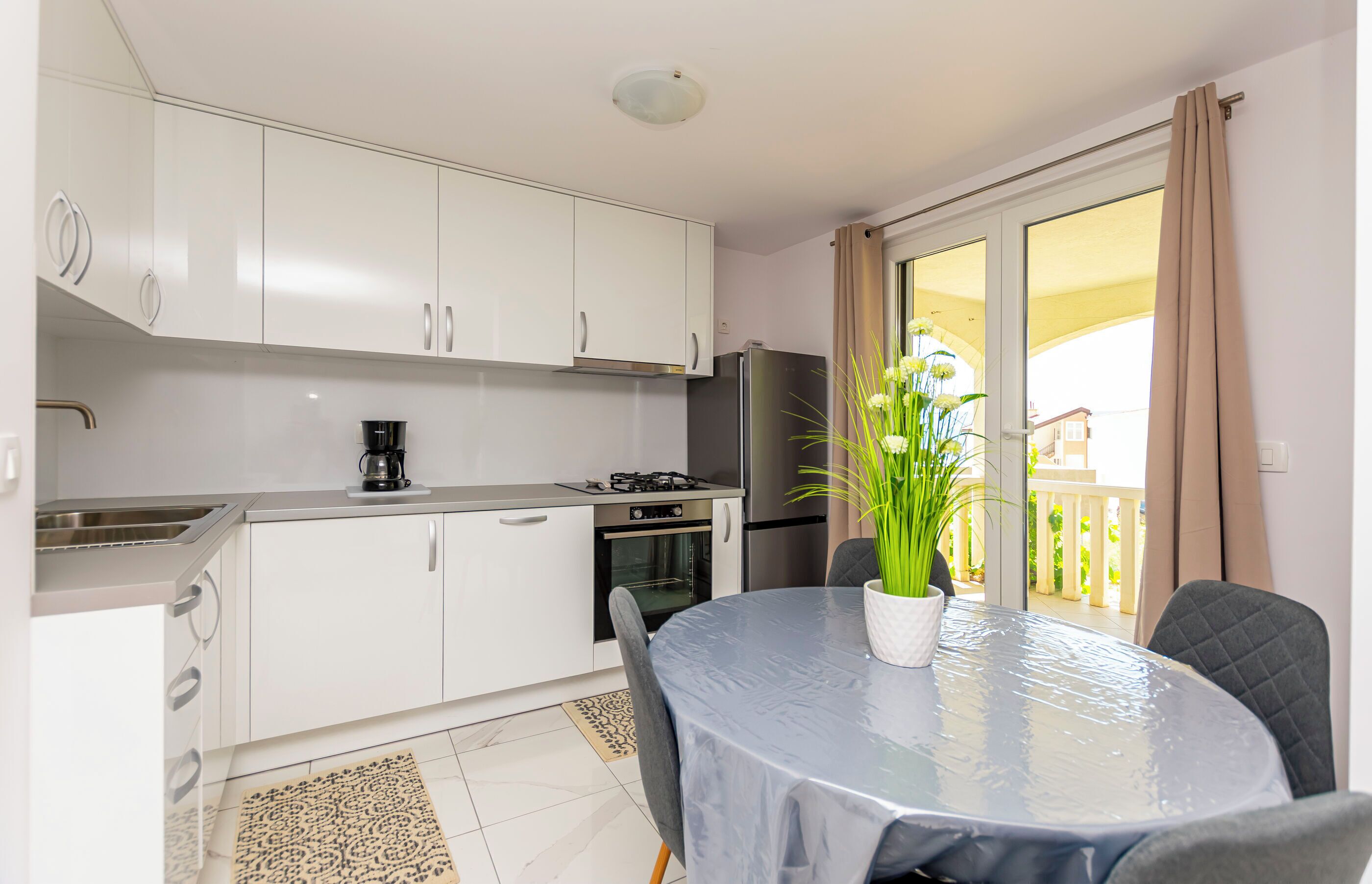Eine moderne Küche mit weißen Schränken, einem Gasherd und einem Kühlschrank. Ein Esstisch steht dort mit einem Topfblumen und einem Stuhl.