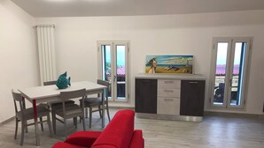 Una sala de estar moderna con un sofá rojo, una mesa de comedor con sillas, una zona de cocina con armarios y un cuadro, y ventanas grandes con vistas al exterior.