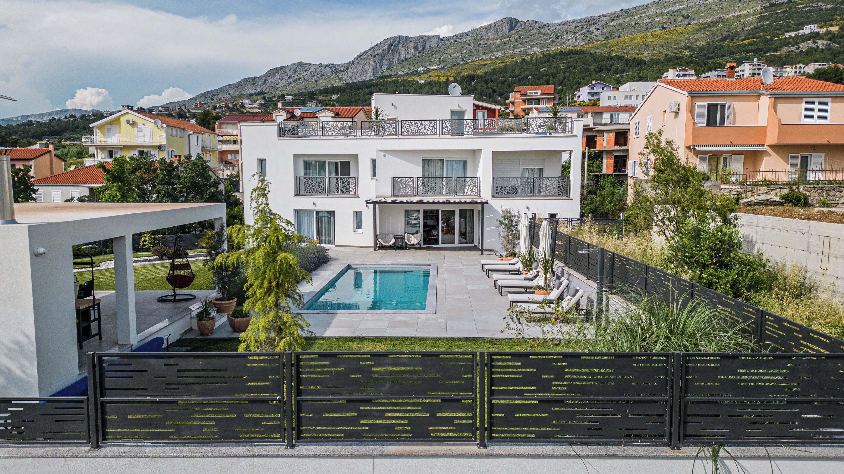 Une maison moderne à deux étages avec une piscine, entourée d’une clôture et de verdure.