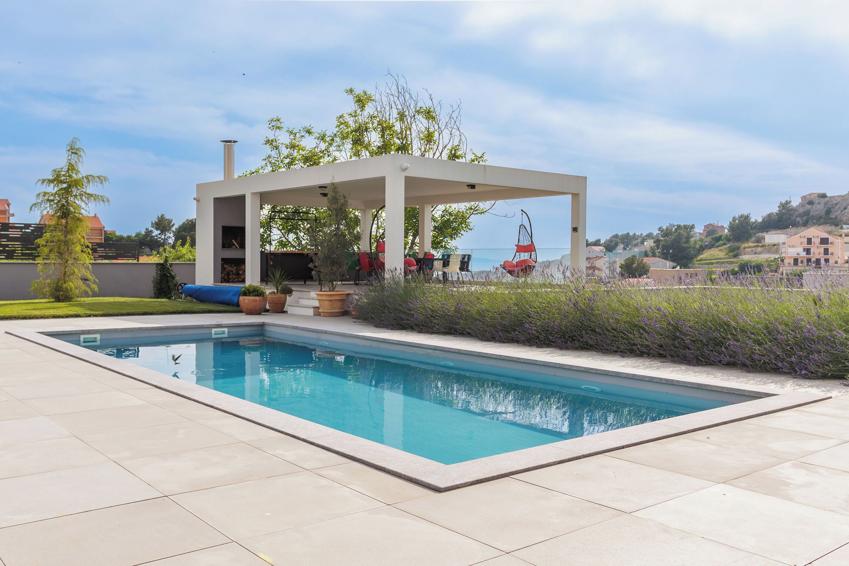 Une piscine rectangulaire agrémentée d’une pergola et d’un espace détente, entourée de plantes de lavande et offrant une vue sur une ville perchée sur une colline.