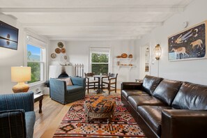 A living room with a sofa, armchair, ottoman, and a dining table with chairs. There is a fireplace, wall decor, and a view through the window. - Tesuque Outpost |3 Casitas |Best views in Santa Fe (Santa Fe)
