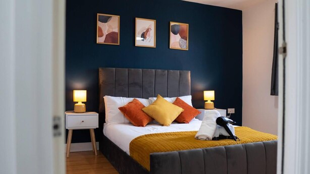 A bedroom with a dark headboard, a bed with white and orange pillows, a mustard-colored blanket, and two bedside lamps. - Stunning 6-bed House in Milton Keynes (Milton Keynes)