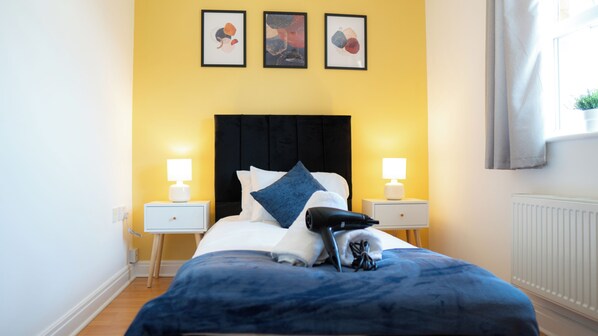 Une chambre à coucher avec un lit, deux tables de chevet avec des lampes, un radiateur et trois œuvres d’art encadrées accrochées au mur.