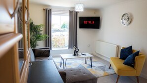 A modern living room with a sofa, a yellow armchair, a coffee table, and a TV displaying "NETFLIX". - Stunning 6-bed House in Milton Keynes (Milton Keynes)