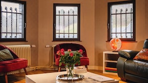 Een woonkamer met een open haard, een rode bank, een zwarte leren bank, een bijzettafeltje met een bloevas en twee ramen met metalen tralies.