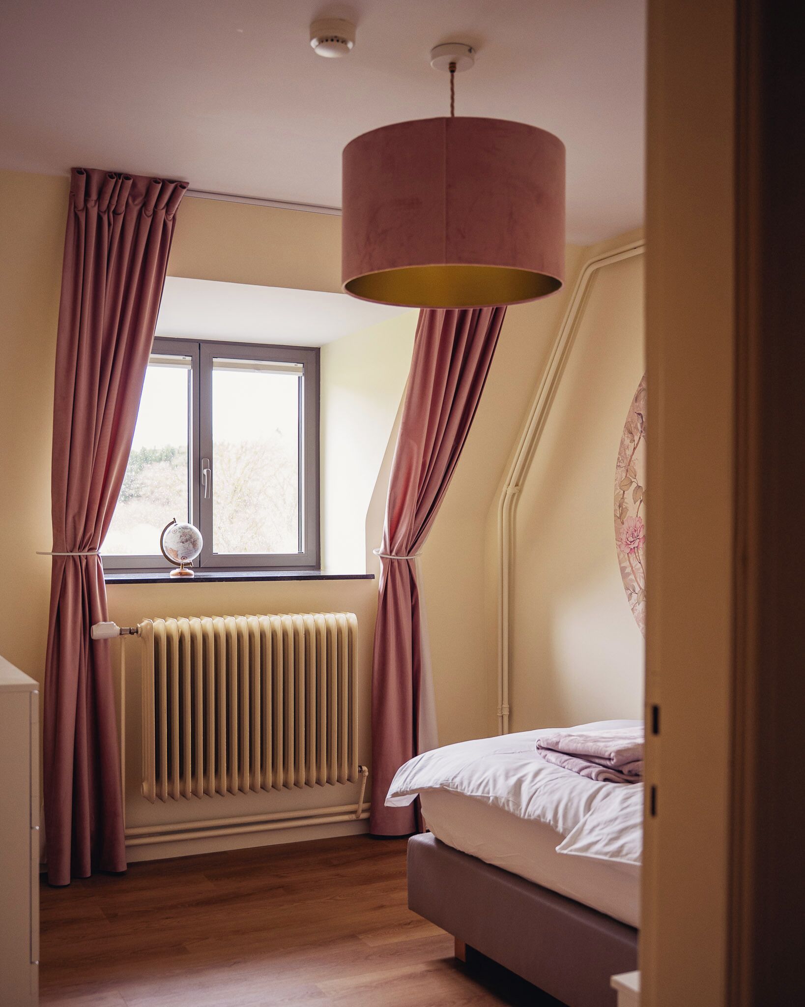 Une chambre à coucher avec un lit, une fenêtre avec des rideaux, un radiateur et un luminaire au plafond.