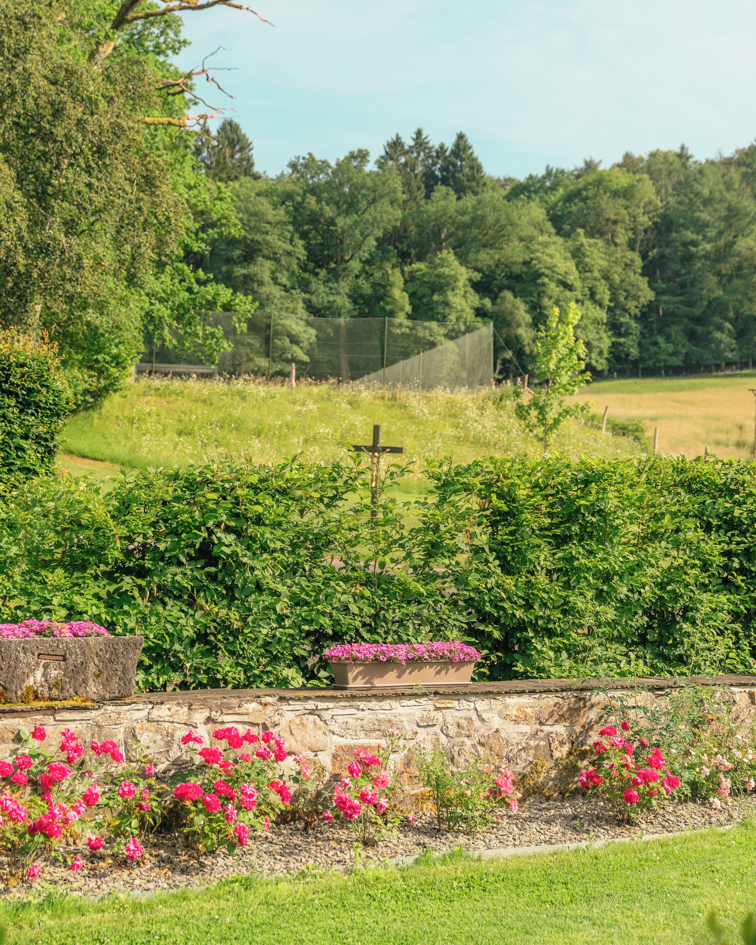 Un mur de pierre surmonté d’une croix, entouré d’une végétation luxuriante et de plantes en fleurs.