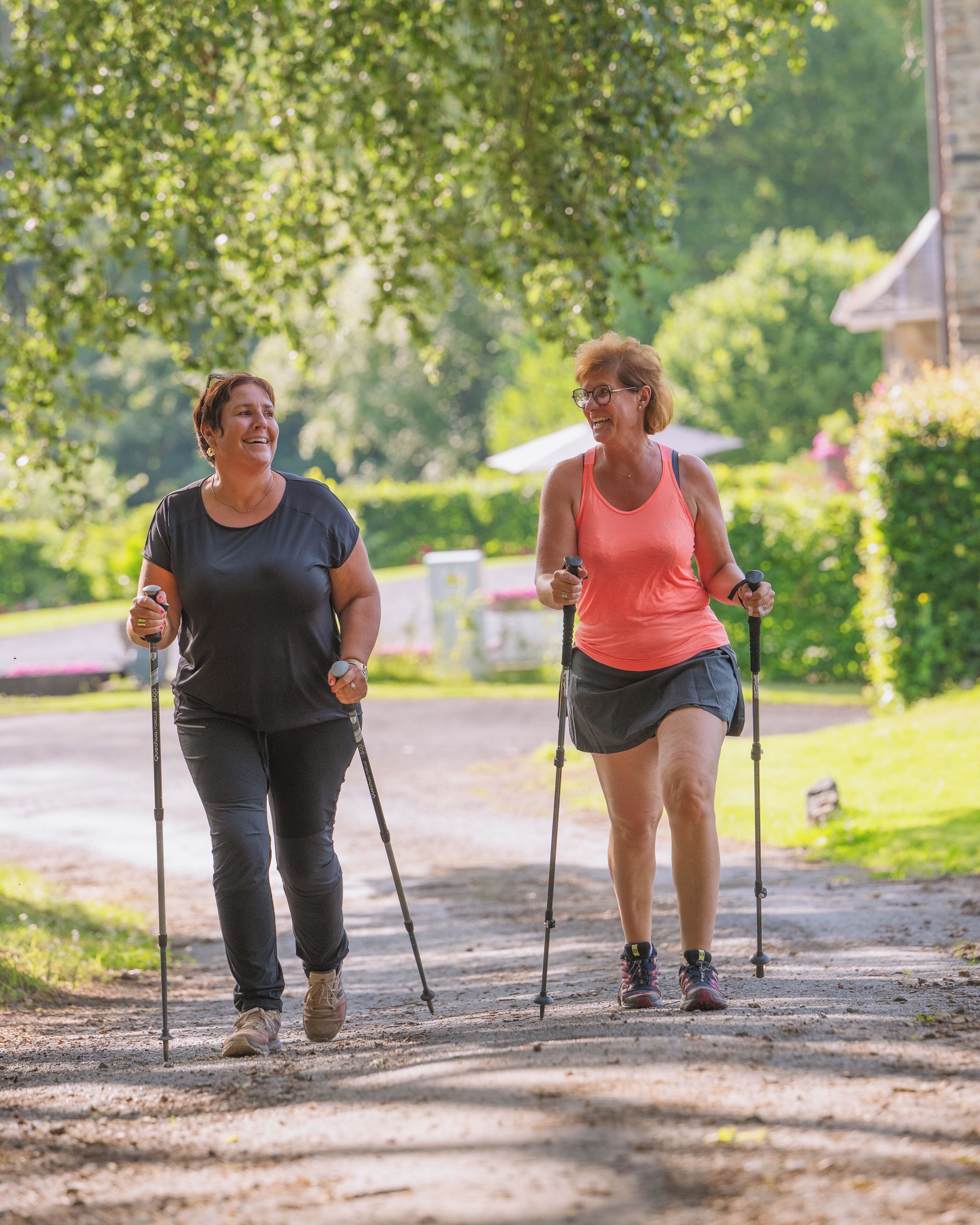 Two individuals walking on a dirt path with walking sticks, surrounded by greenery and a building in the background.