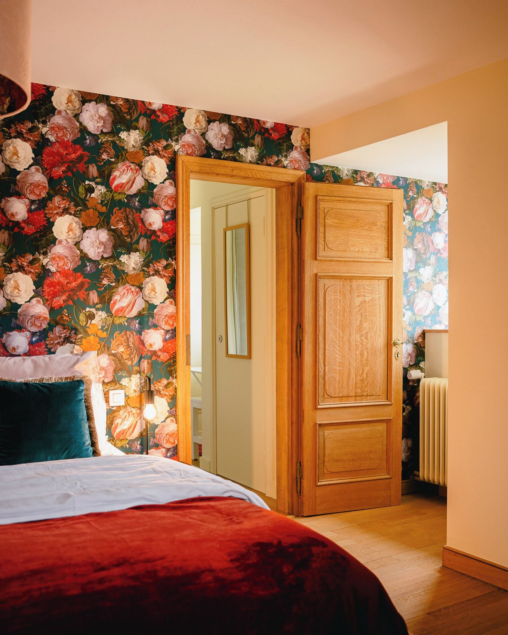 Un dormitorio con papel tapiz floral, una puerta de madera, una cama con manta roja y una mesita de noche con lámpara.