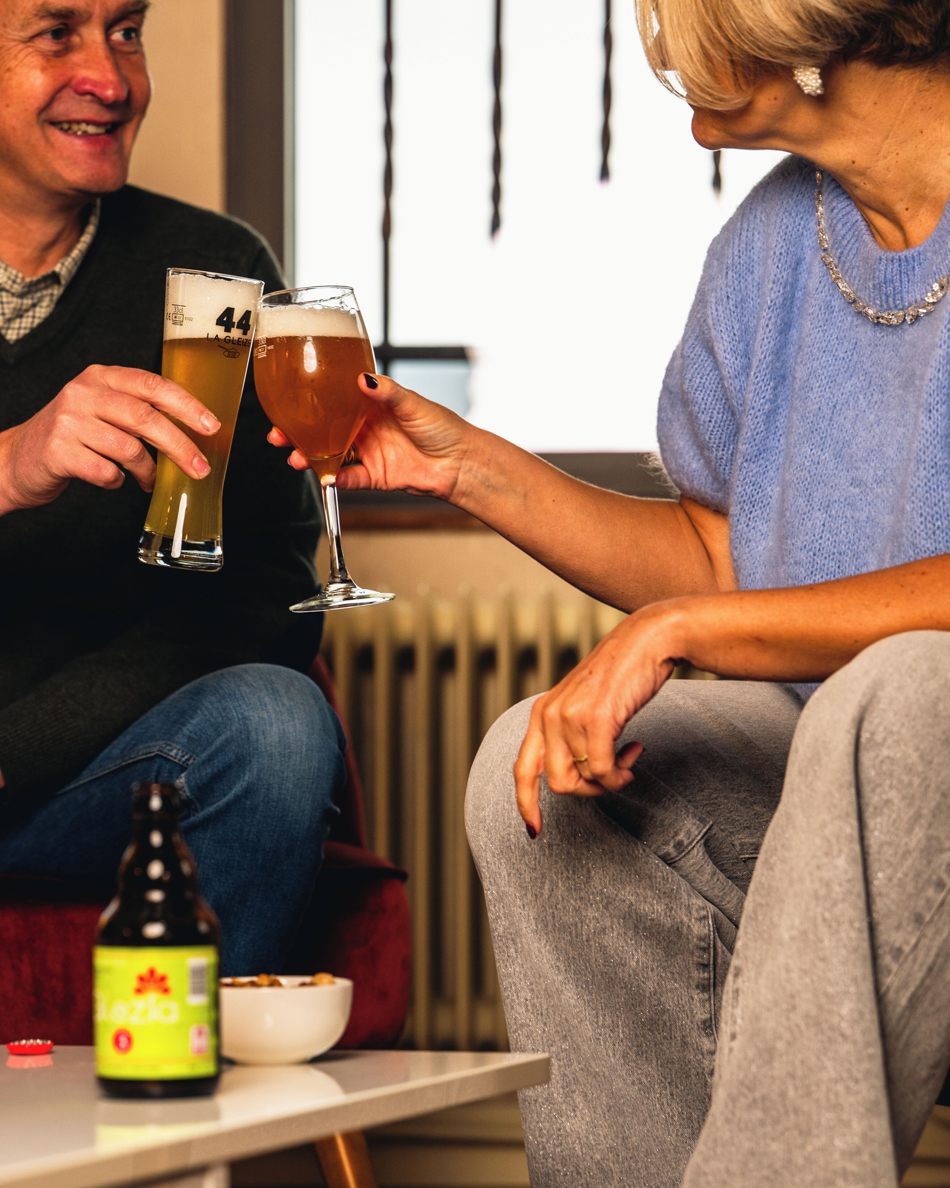 Dos personas brindando con vasos de cerveza, una botella de cerveza y un bol sobre la mesa.