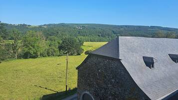 A stone building with a sloped roof, a gated entrance, and a view of a green field and forested hills in the background.
