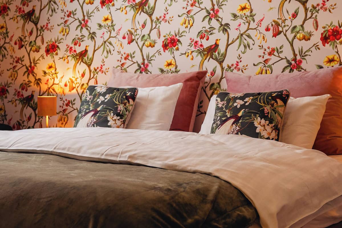 A bedroom with floral wallpaper, a bed with white linens, and a bedside lamp.