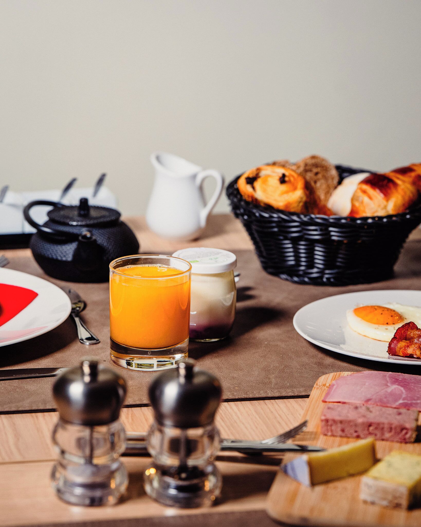 Una mesa de desayuno con un vaso de zumo de naranja, una jarra de leche, una cesta de cruasanes y un plato con huevos y jamón.