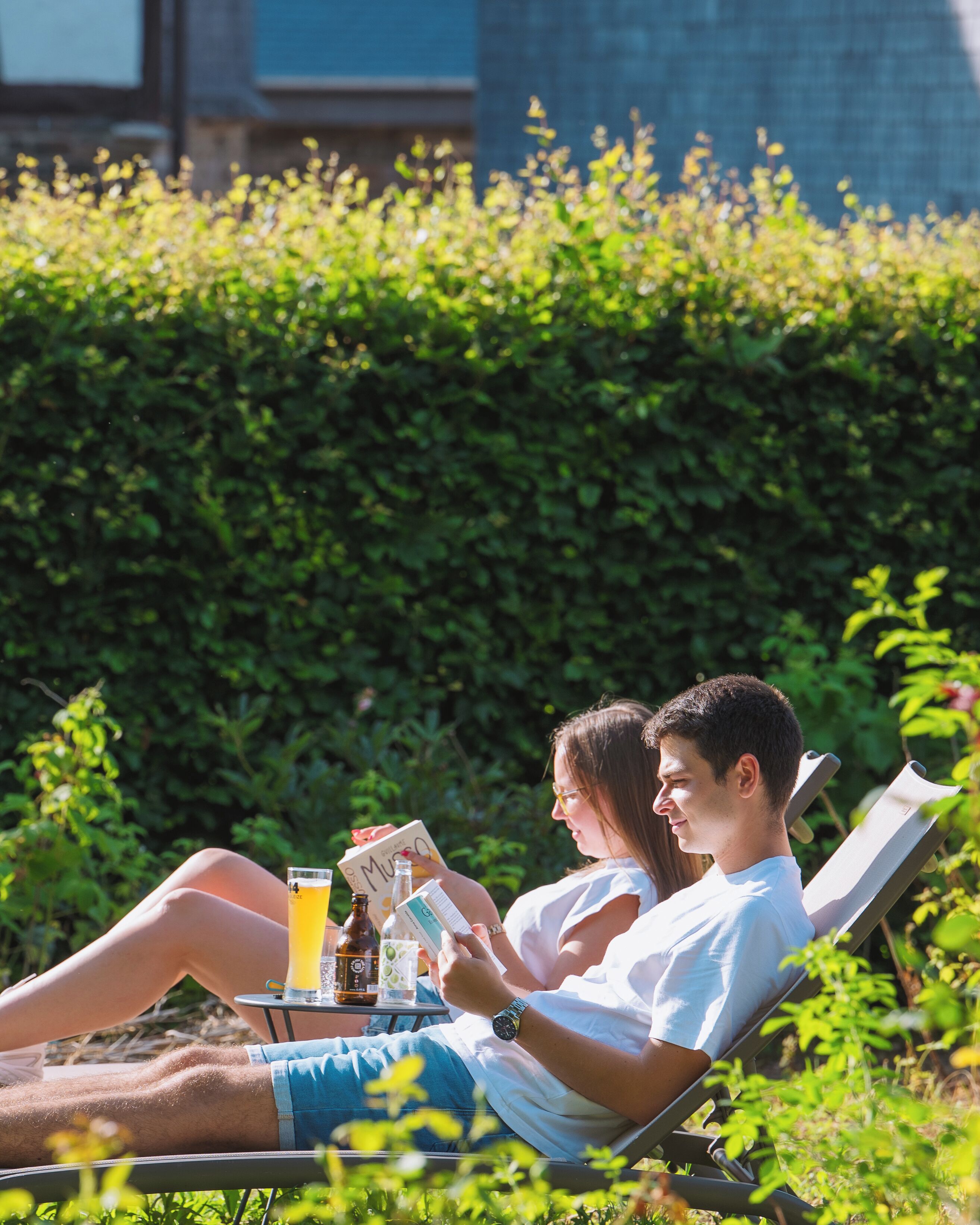 A couple relaxing on a sun lounger with drinks and a book, surrounded by greenery.