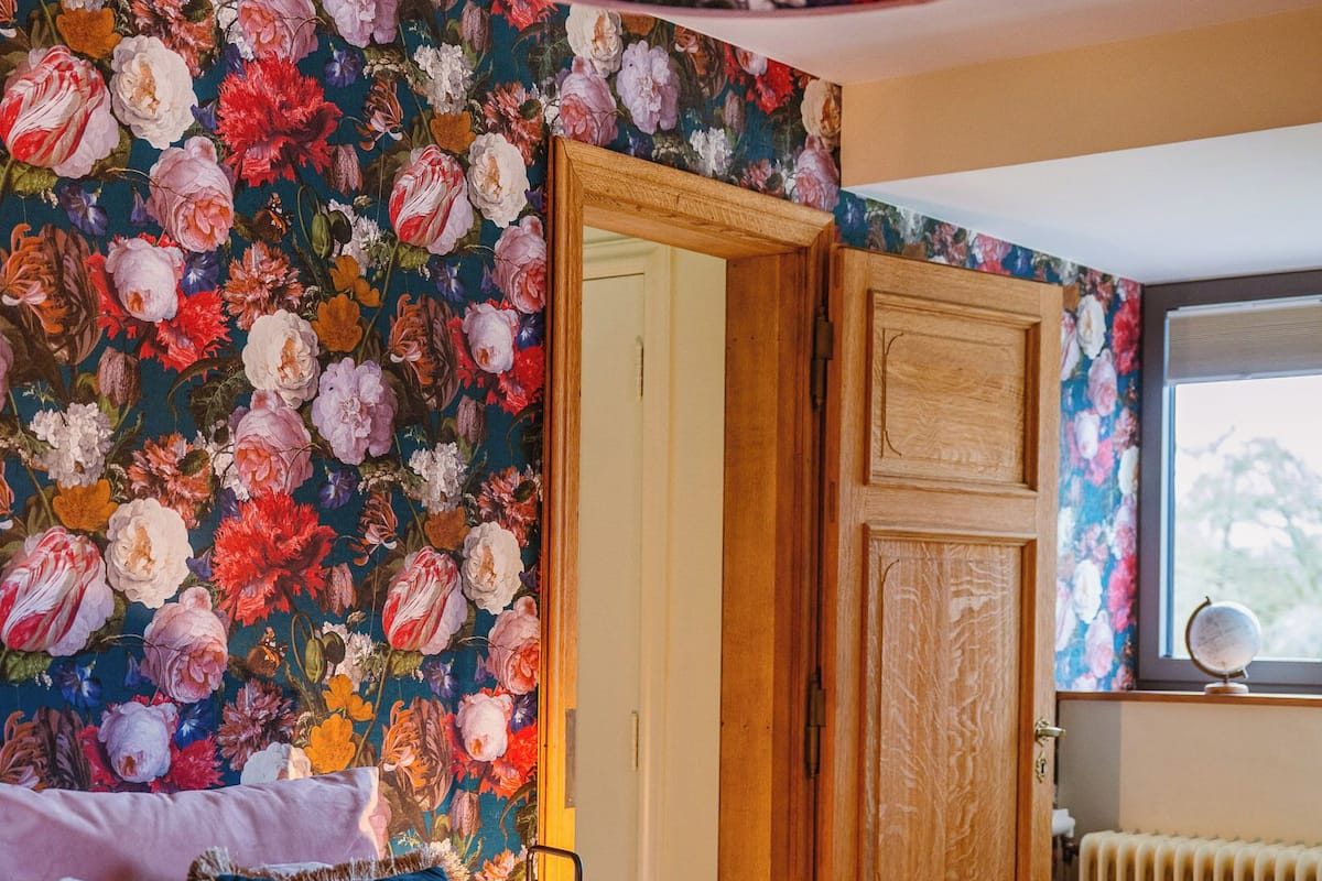 A bedroom with a floral wallpaper, a wooden door, a bed with pillows, a bedside lamp, and a window with a view.