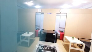 Ein Zimmer mit weißem Schreibtisch, einem kleinen Tisch mit Pflanze, einem schwarzen Schrank und einem roten Tisch mit diversen Gegenständen.