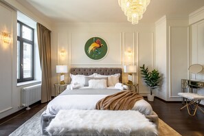 Makuuhuoneessa on suuri sänky, kaksi yöpöytää, kattokruunu, ikkuna, jossa on verhot, ja seinällä oleva taulu.